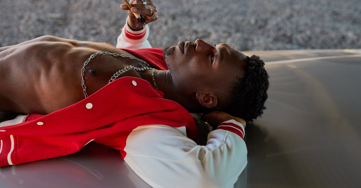 A young man in a red jacket relaxes atop a car hood, smoking and contemplating the sky.