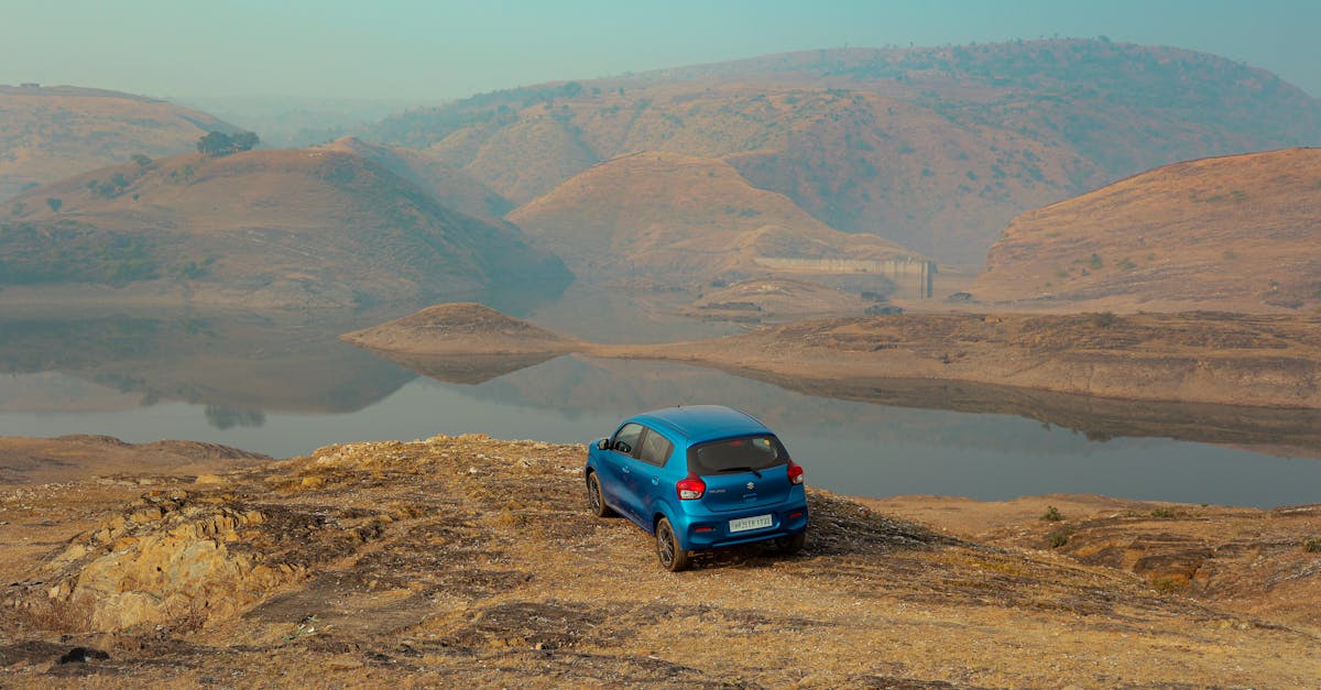 A blue car parked on a cliff with a panoramic view of a tranquil lake and surrounding hills under a clear sky.