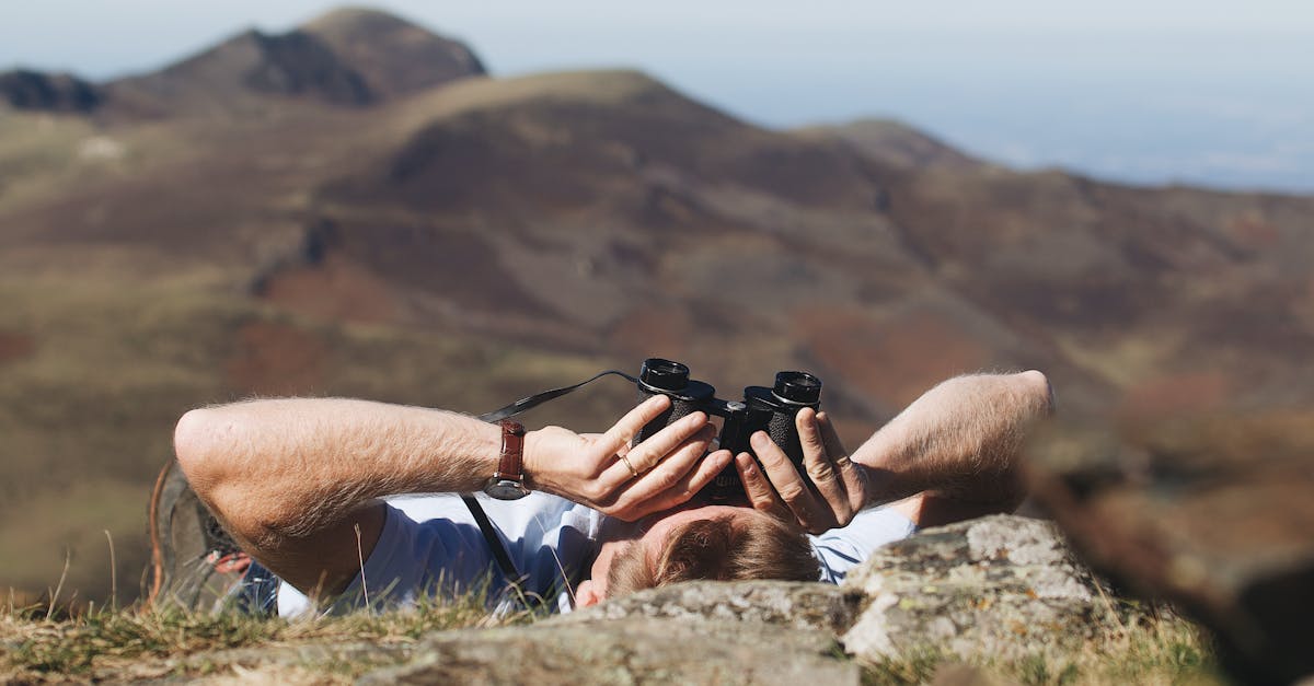 A man lies on a mountainside observing the landscape through binoculars on a clear day.