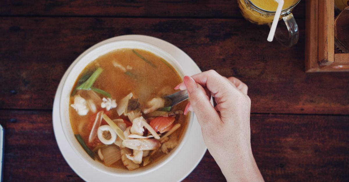 A close-up view of a person enjoying a bowl of seafood soup with a cold beverage on a wooden table.