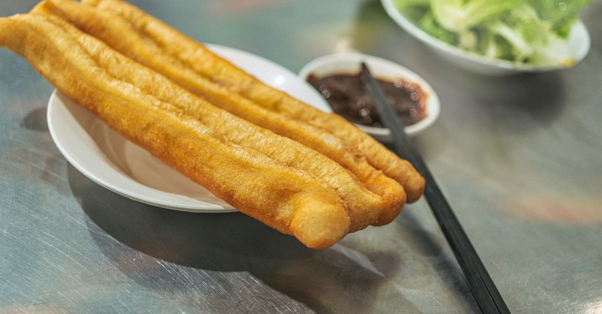 Close-up of crispy youtiao served with dipping sauce and chopsticks on a metal table.
