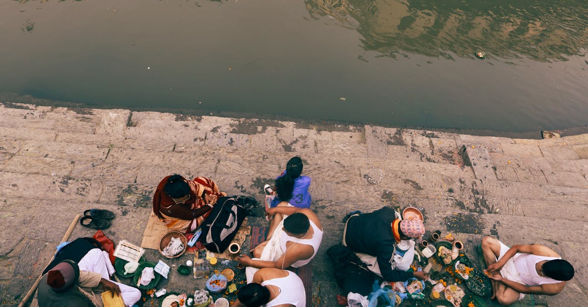 Group of adults sitting by a riverbank sharing a meal in a traditional setting.