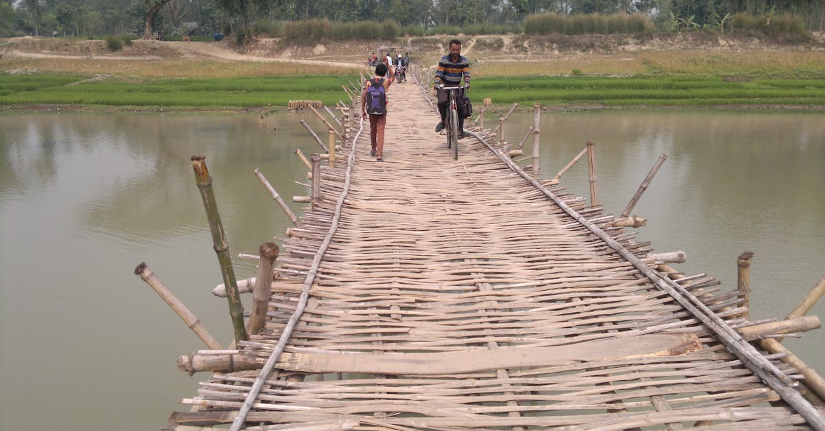 A rustic bamboo footbridge crossing a calm river with people walking and a man bicycling.