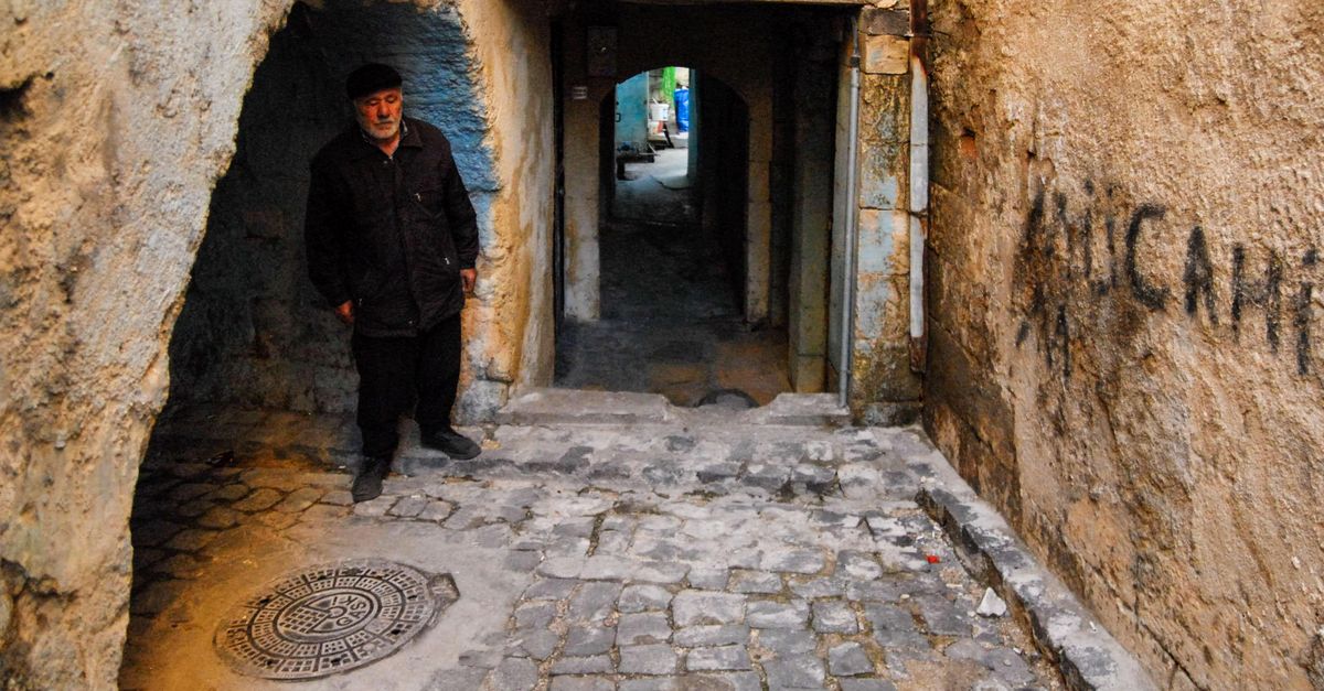 An elderly man stands in a cobblestone passageway with stone walls and graffiti. Vandalized signs add character to the scene.