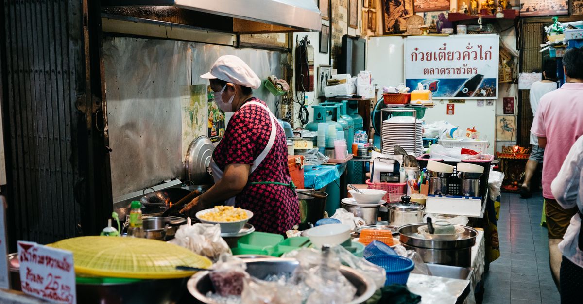 Street food vendor in a bustling Bangkok market kitchen preparing dishes with fresh ingredients.