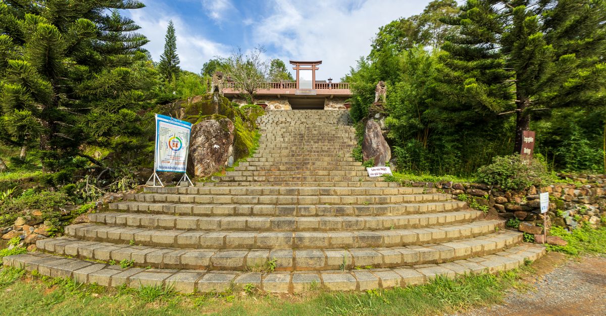 Wide stone steps lead to a traditional temple gate surrounded by lush greenery in an outdoor setting.