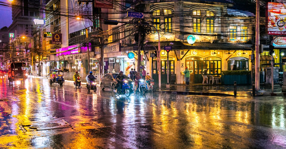 Explore the vibrant nightlife of Bangkok on a rainy evening, with bustling streets and neon lights reflecting in water.