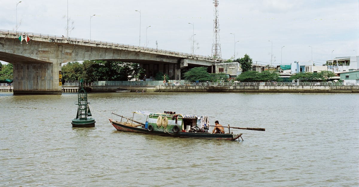 A man navigates a small boat under a concrete bridge on a city river, with a buoy and distant buildings.
