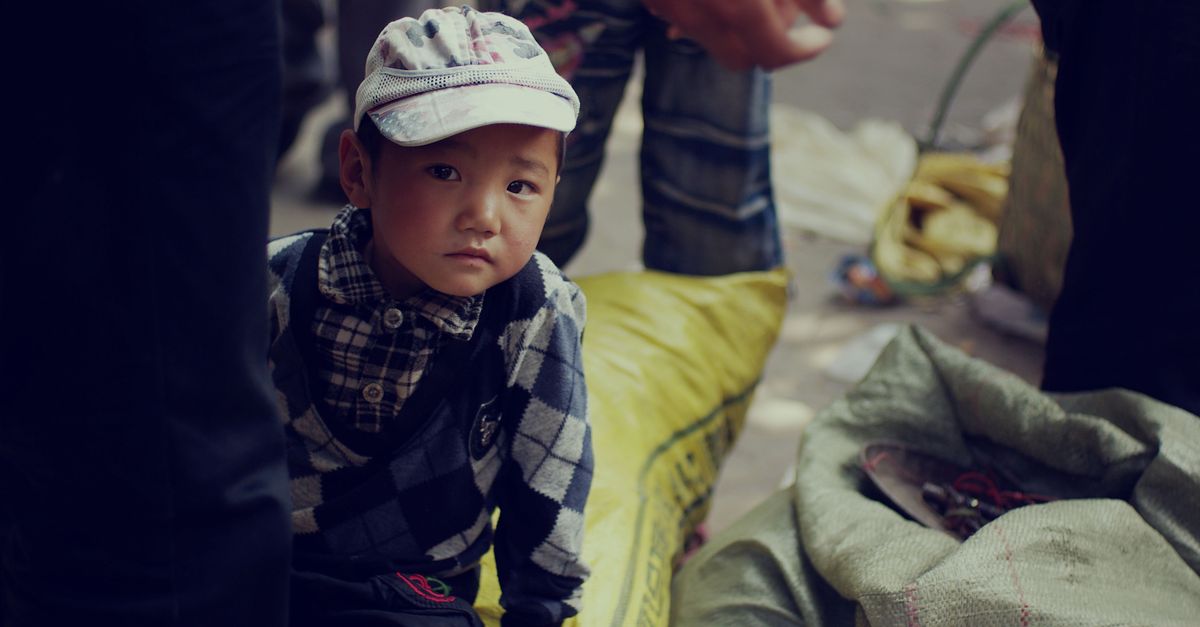 A young child wearing a cap gazes thoughtfully amidst an outdoor market setting, surrounded by sacks.