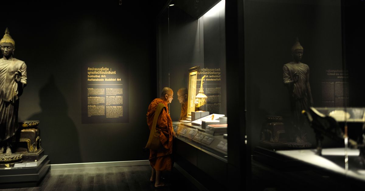 Monk in religious attire observing Buddhist artifacts in dimly lit museum.