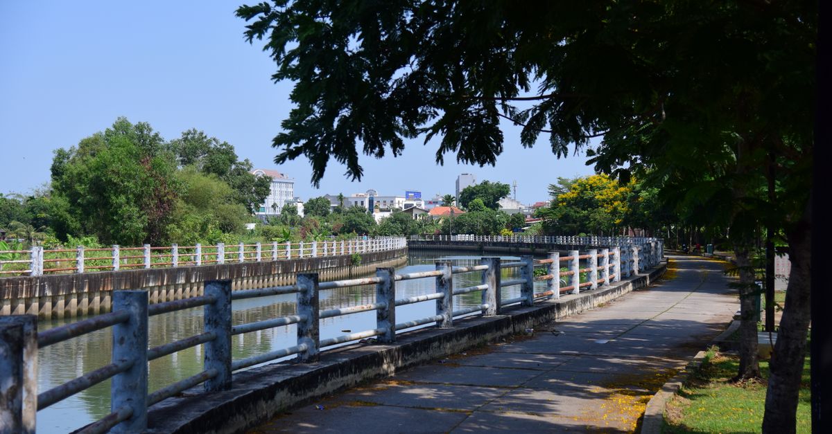 A serene riverside park path with trees and railings under a blue sky.