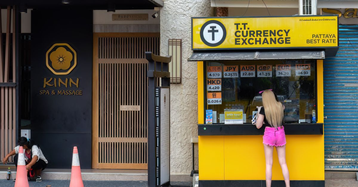 Woman at currency exchange booth in Pattaya, Thailand, with currency rates displayed.
