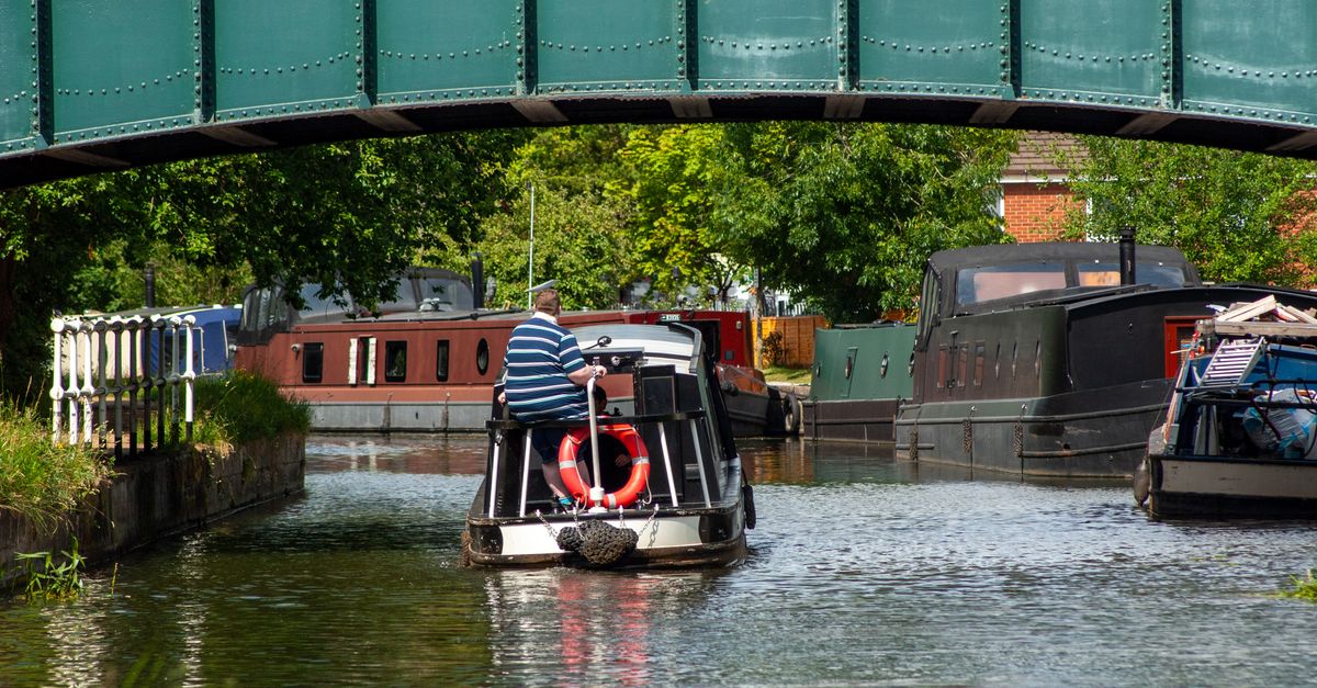 A canal boat navigates under a green bridge on a sunny day with lush surroundings and other boats docked nearby.