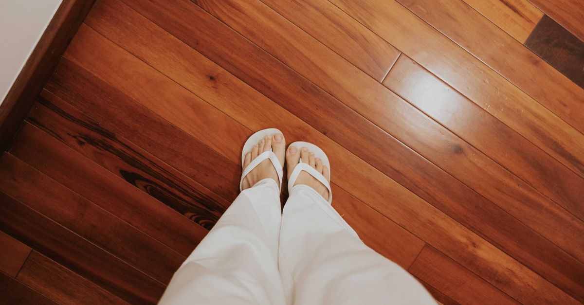 View of person wearing flip-flops and white pants standing on a wooden floor.