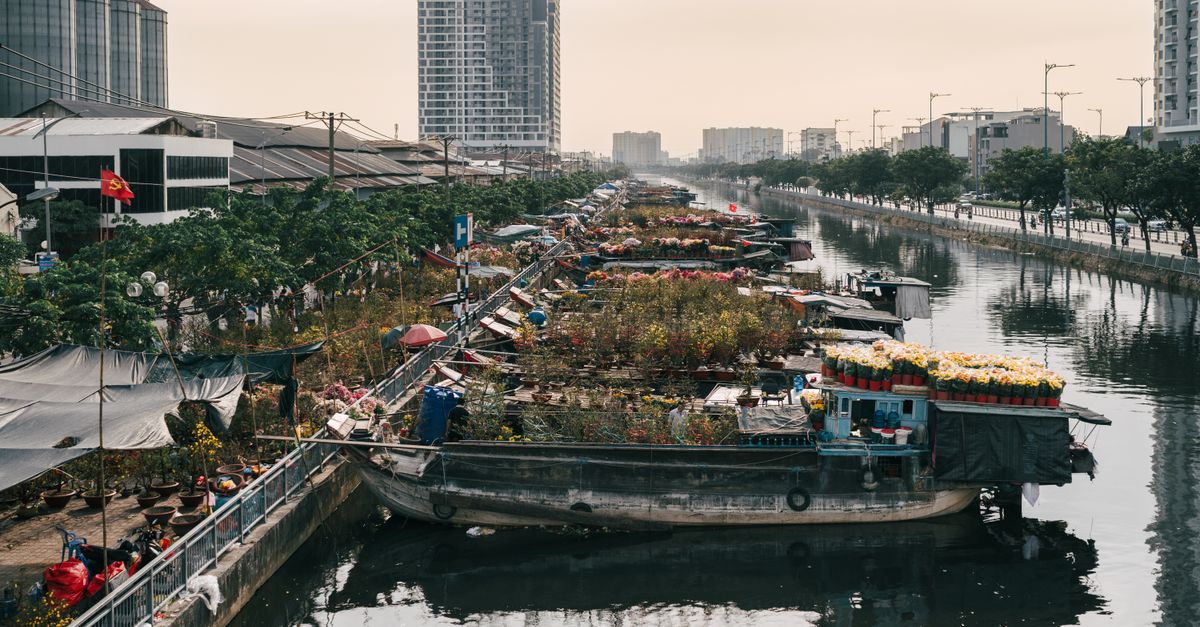 Flower boats line a calm river in a bustling city with towering buildings.