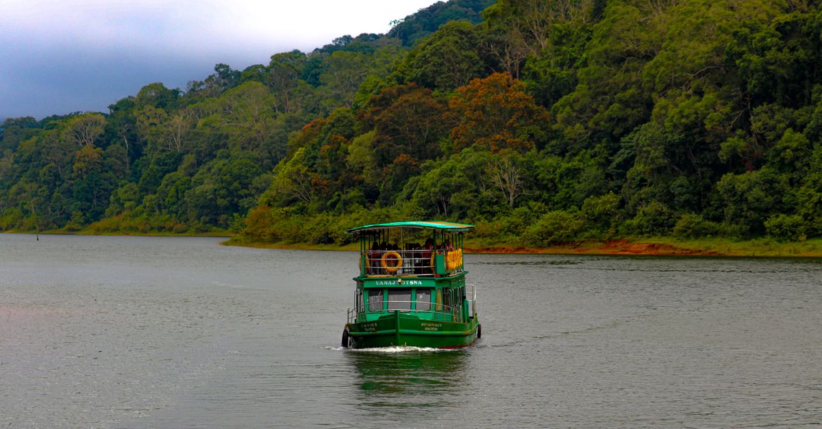 A green boat navigates a tranquil lake bordered by lush green forest under a cloudy sky.
