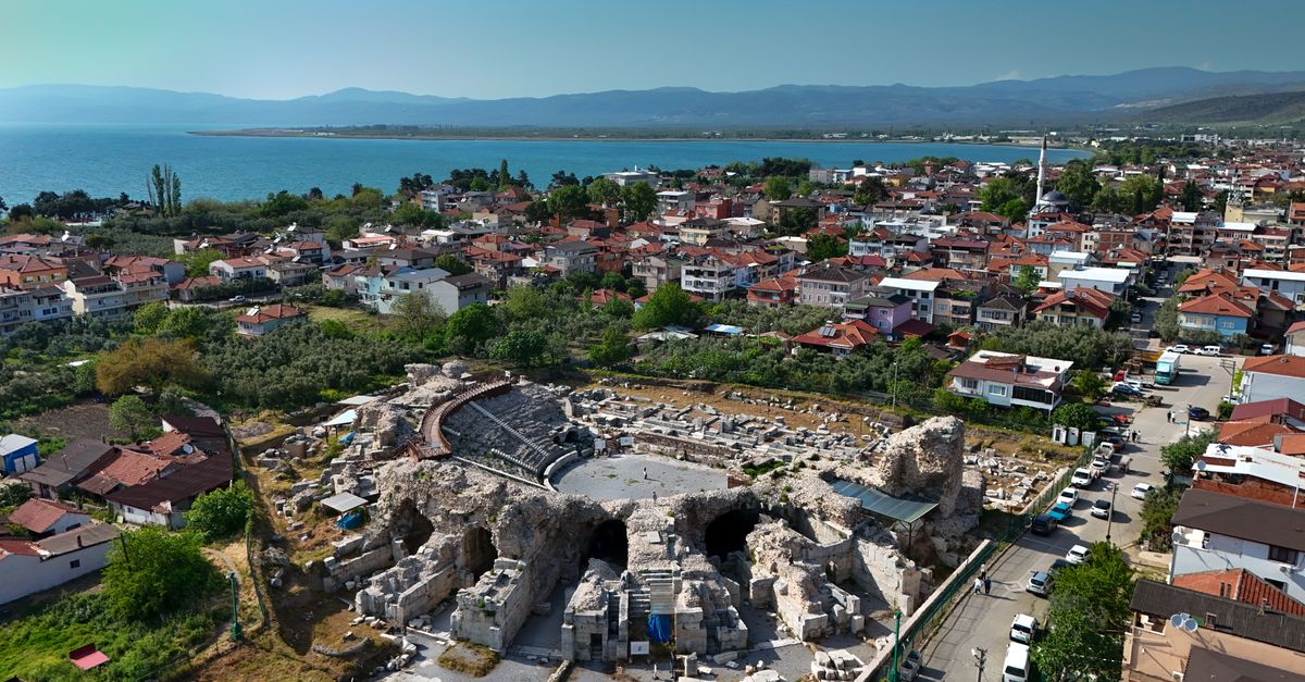 Aerial shot showcasing an ancient amphitheater surrounded by a vibrant coastal town with distant mountains.