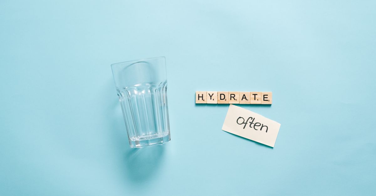 A minimalist composition of a glass with Scrabble tiles spelling 'HYDRATE' on a blue background.