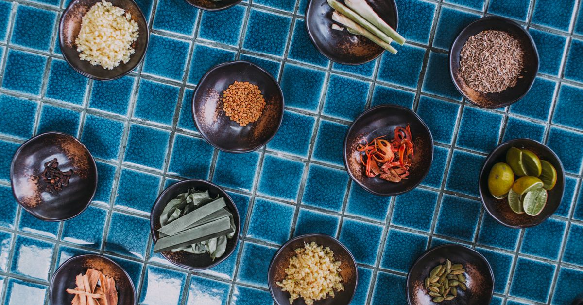 Assorted spices and ingredients in bowls arranged on blue tiles, top-down view.