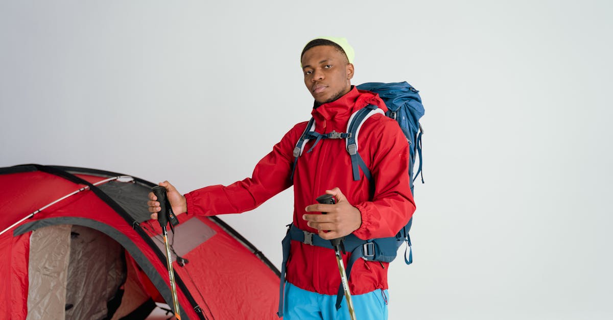 Male hiker in red jacket with backpack and ski poles beside a tent, ready for adventure.
