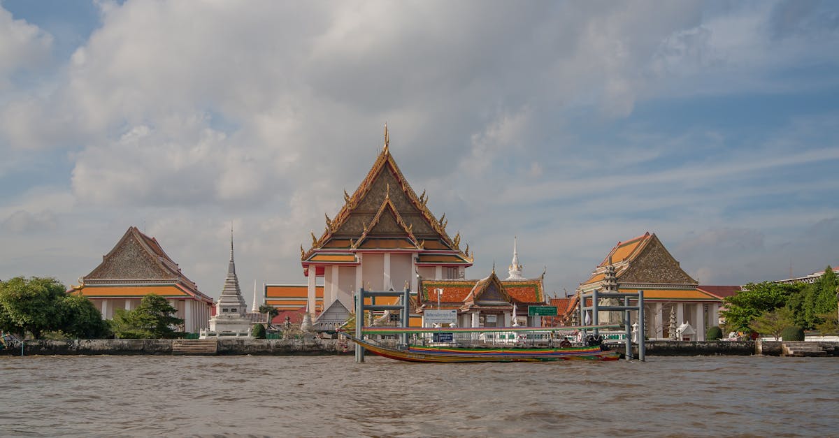 Scenic view of a colorful temple by the river in Bangkok under a partly cloudy sky.