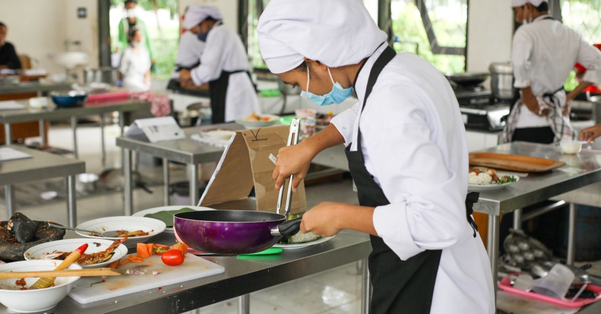 Culinary students in uniforms practice cooking skills in a professional kitchen setting.