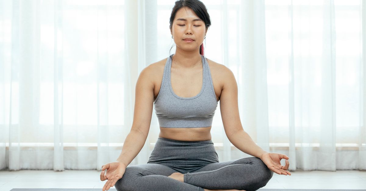 Asian woman meditating indoors on yoga mat, promoting fitness and wellness.