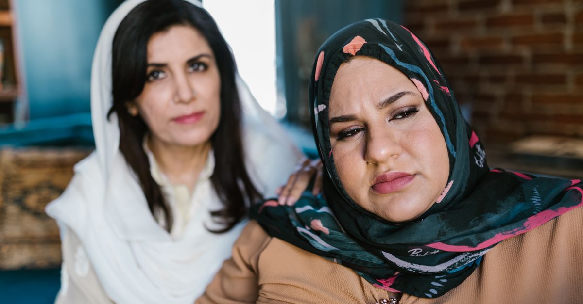Two middle-aged women in hijabs indoors, sharing emotional support. One has their hand on the other's shoulder.