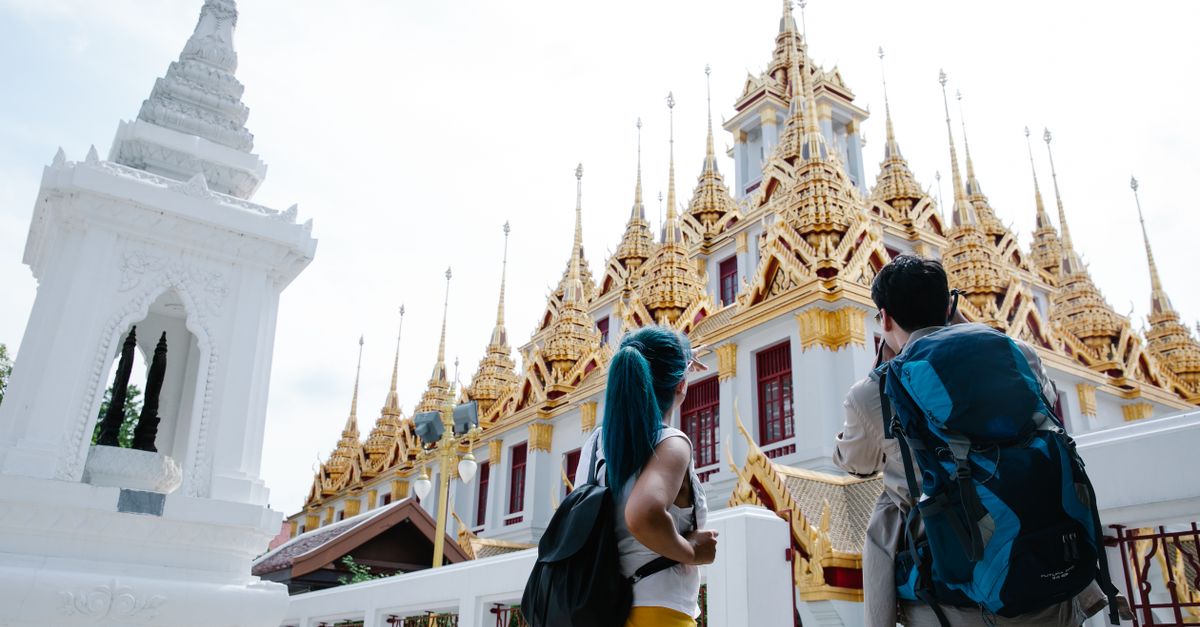 Two tourists with backpacks admire the ornate facade of a traditional Thai temple.
