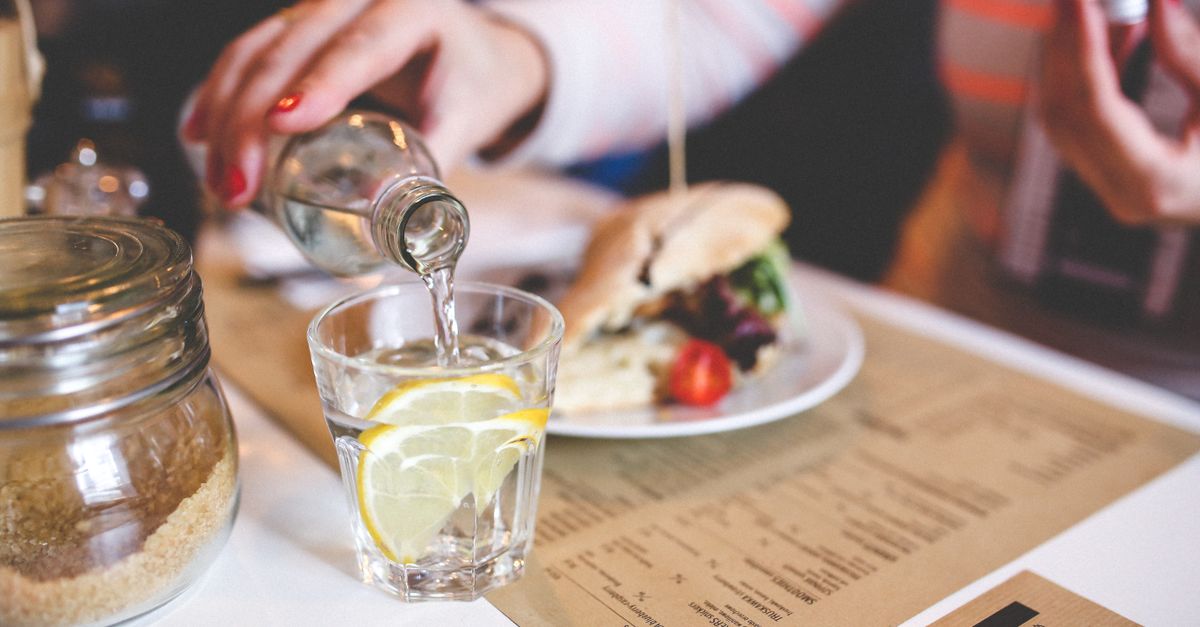 Close-up of water being poured into a glass with lemon on a restaurant table, next to a sandwich and sugar jar.